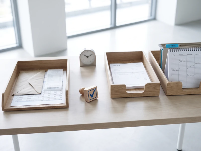 A desk holds paper trays with an invoice, an envelope, a checkmark stamp, and a wall calendar.